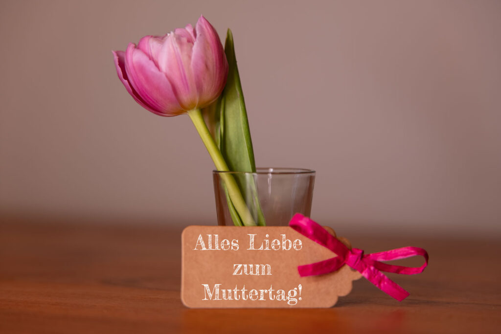 Eine frische rosa Tulpe in einem Glas mit einem Geschenkanhänger und der Aufschrift Alles Liebe zum Muttertag!