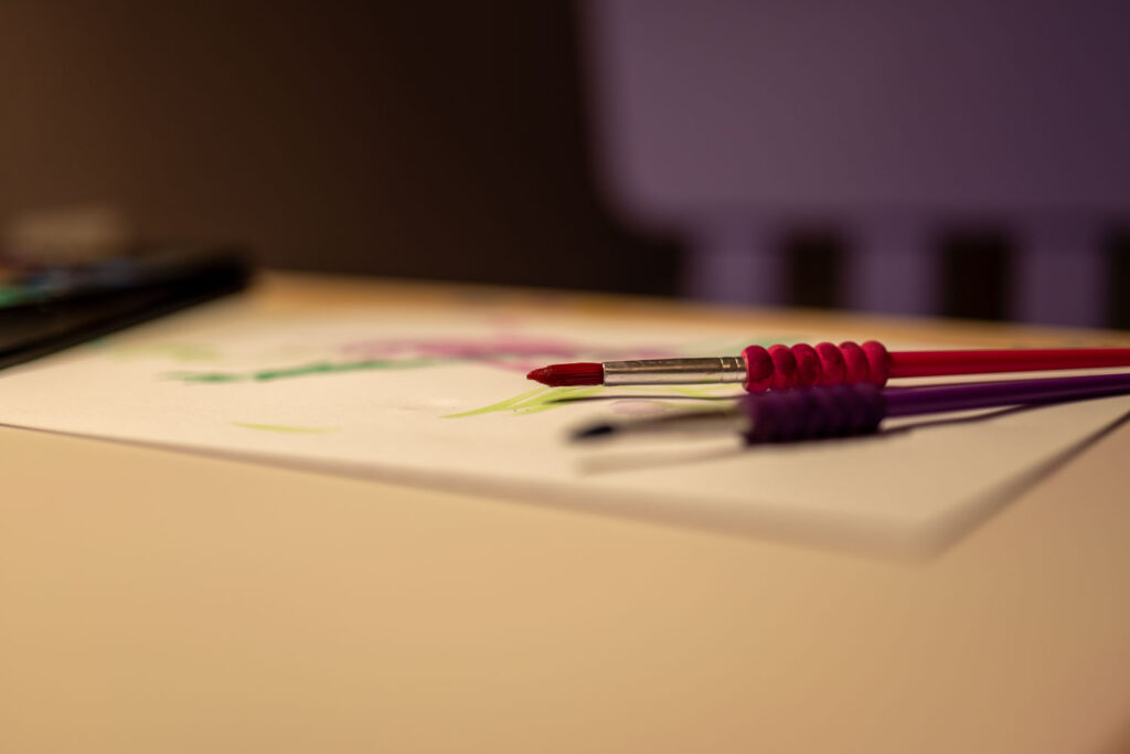 Zwei dicke, bunte Pinsel in Pink und Lila liegen auf einem Blatt Papier mit ersten Wasserfarben-Spuren bereit zum Malen.