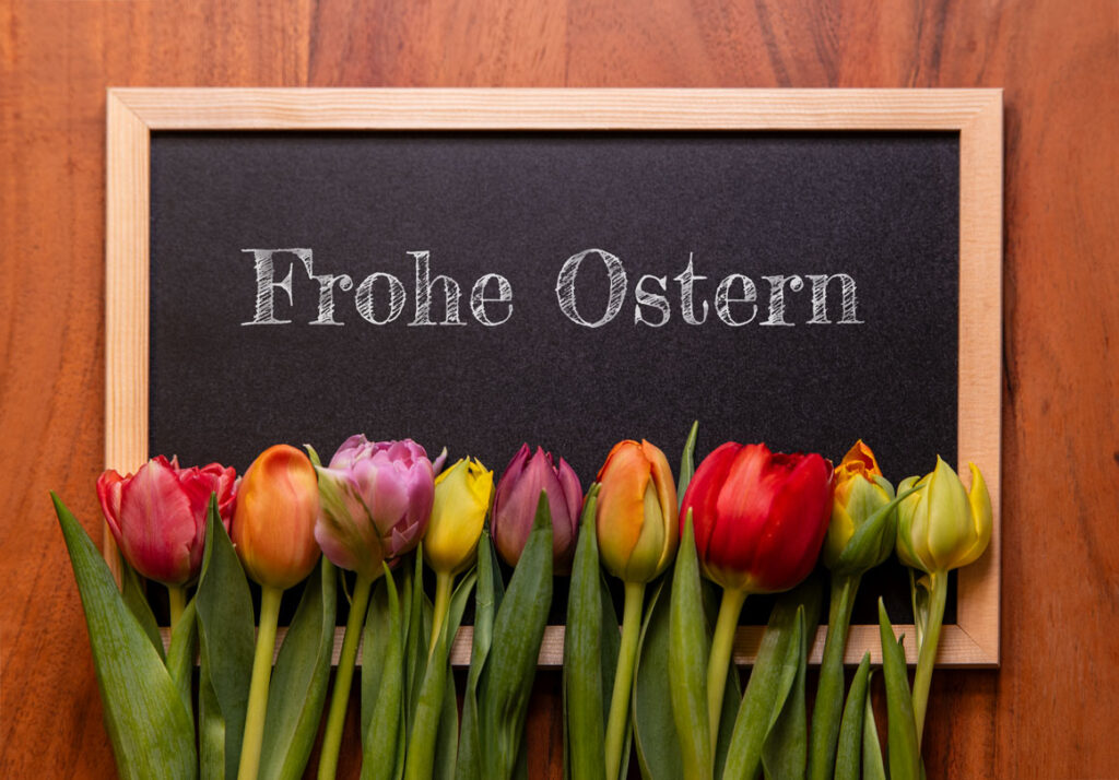 Eine schwarze Kreidetafel mit der Aufschrift Frohe Ostern, davor liegen bunte frische Tulpen auf einem Holztisch.