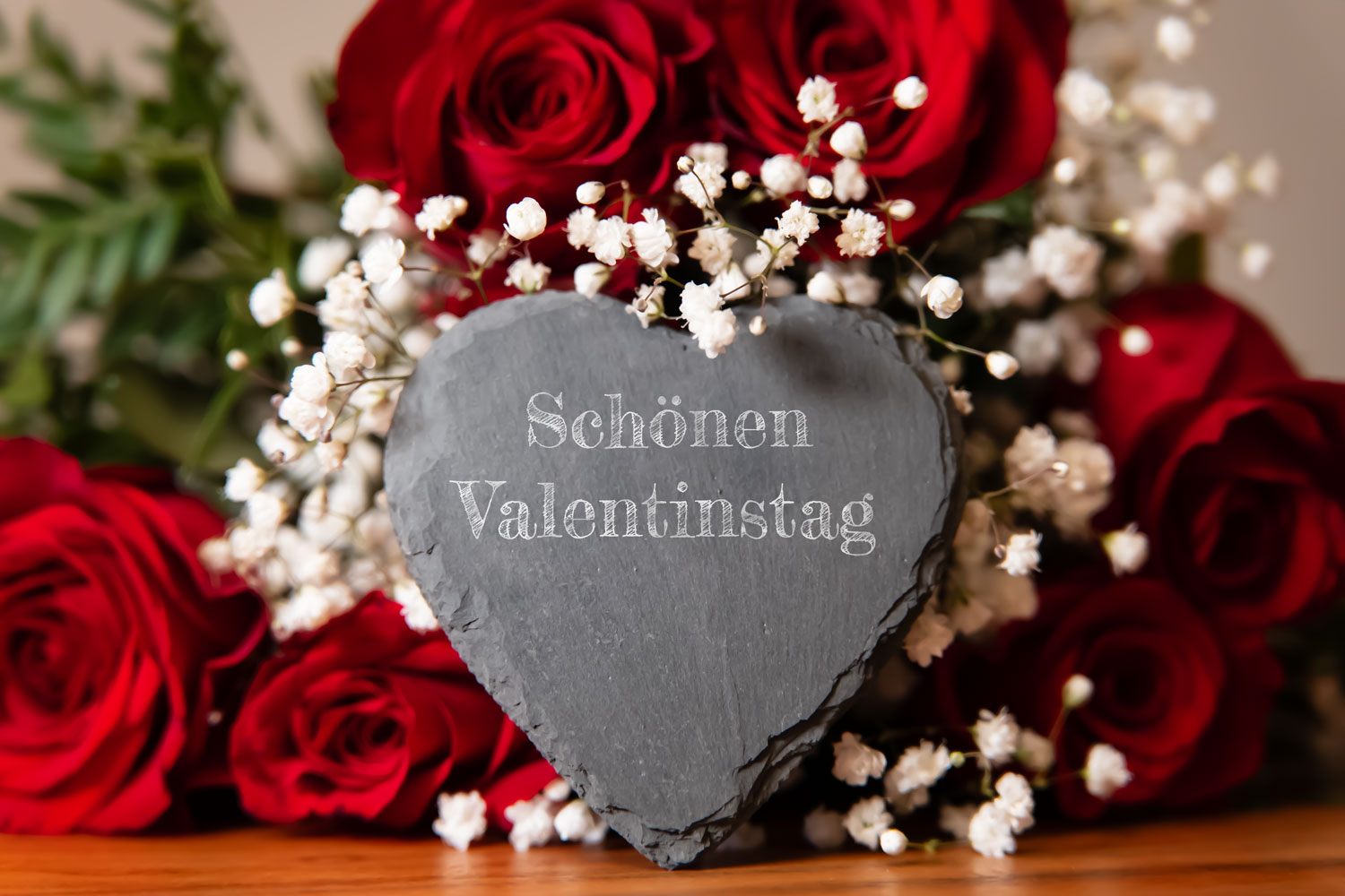 Ein herzförmiger Schieferstein mit der Aufschrift Schönen Valentinstag, umgeben von roten Rosen und weißem Schleierkraut