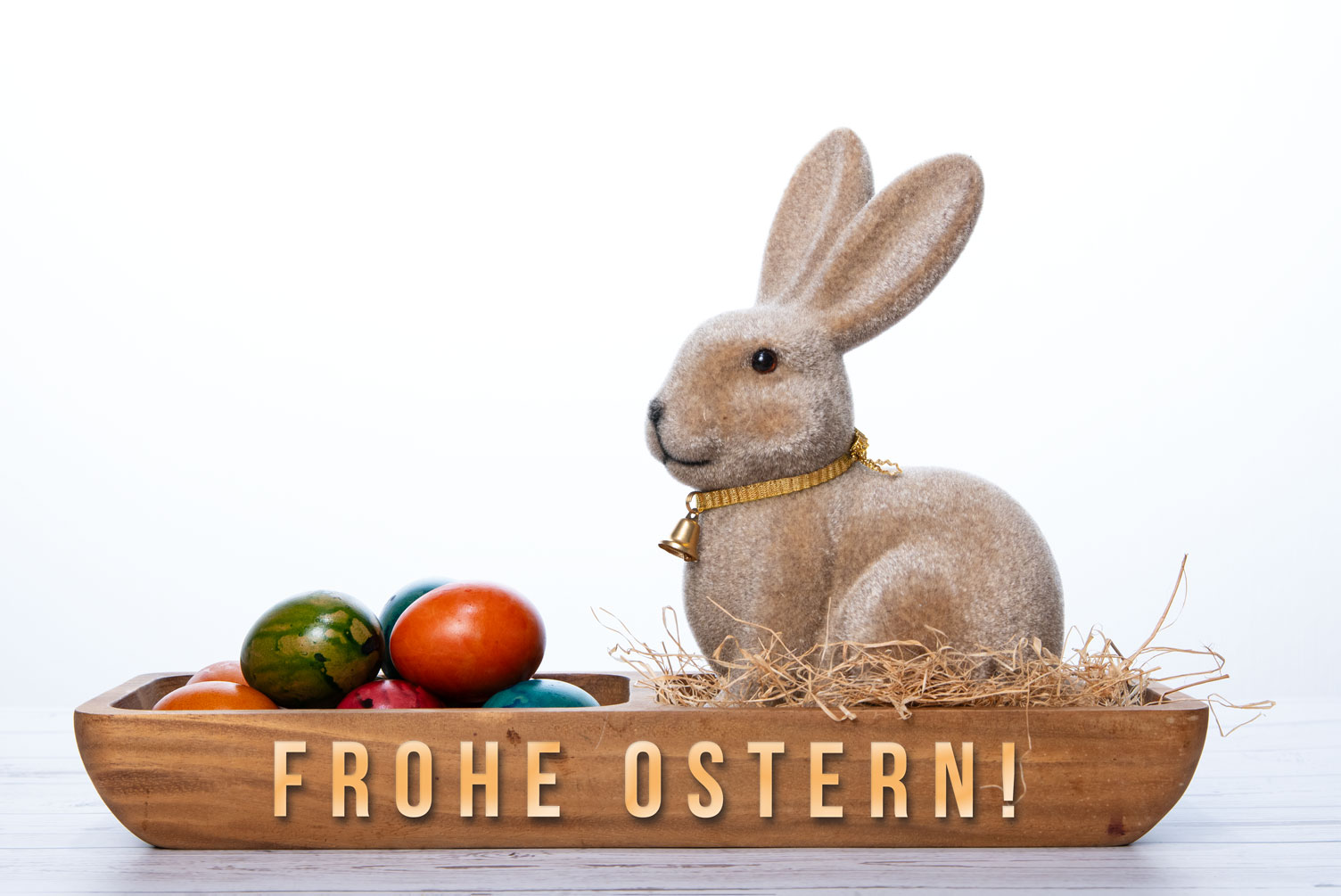 Ein flauschiger Osterhase mit Glocke sitzt in einer Holzschale mit bunten Ostereiern und dem goldenen Text "FROHE OSTERN!".