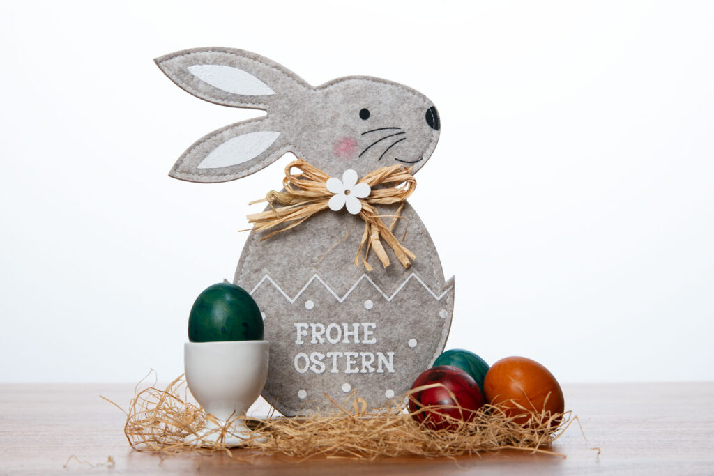Ein Filzhase mit der Aufschrift "Frohe Ostern" steht neben bunten Ostereiern und einem weißen Eierbecher auf etwas Stroh.