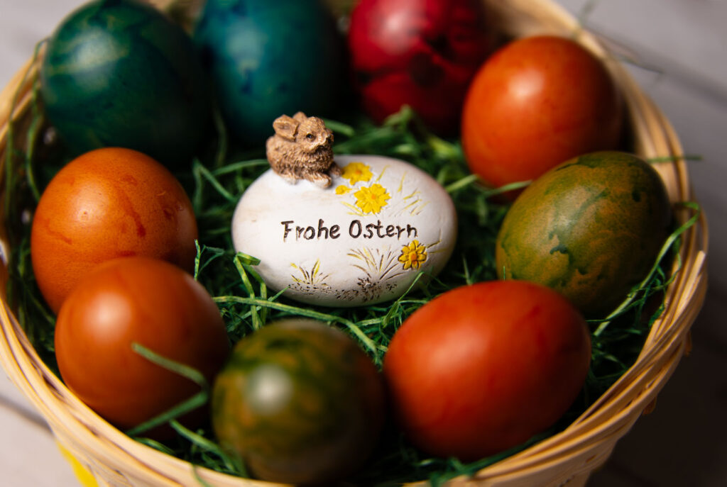 Ein buntes Osternest mit gefärbten Ostereiern und einem weißen Deko-Ei in der Mitte, auf dem "Frohe Ostern" steht und ein kleiner Hase sitzt.