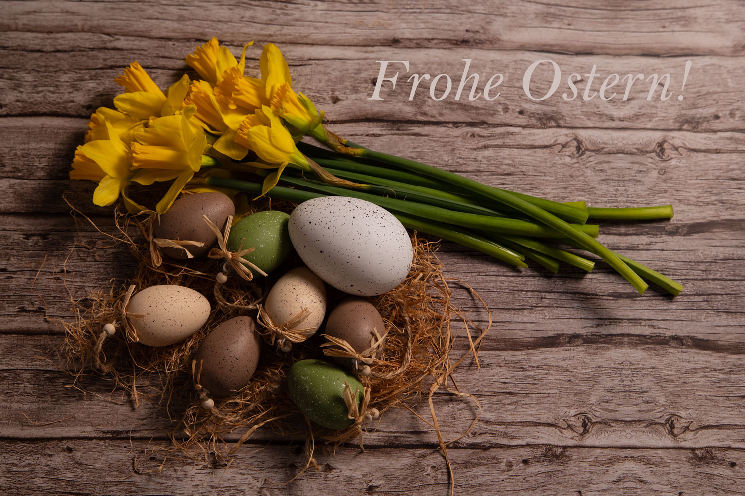 Gesprenkelte Ostereier in einem Strohnest neben frischen gelben Narzissen und dem Schriftzug „Frohe Ostern!“ auf einem rustikalen Holztisch.