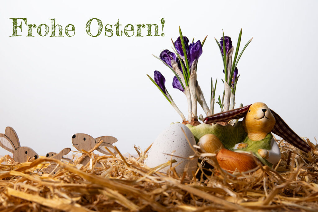 Osterdekoration mit einem Keramik-Hasen, echten lila Krokussen, kleinen Holzhasen und einem Ei im Stroh vor weißem Hintergrund mit dem grünen Text „Frohe Ostern!“.