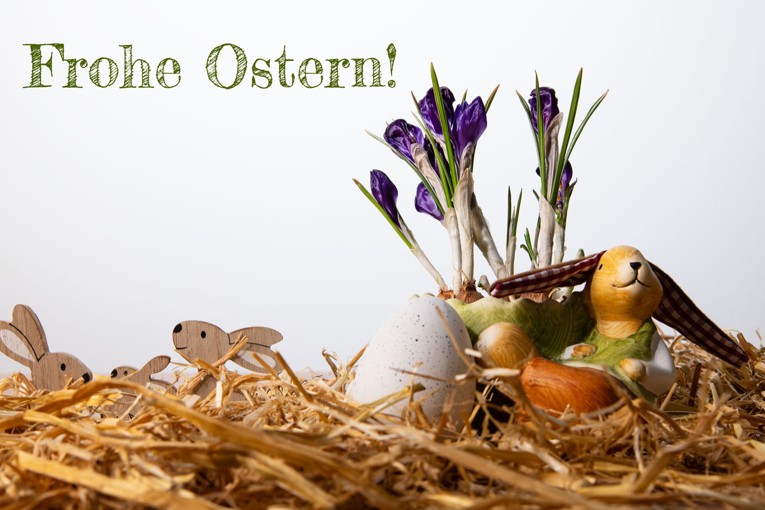 Osterdekoration mit einem Keramik-Hasen, echten lila Krokussen, kleinen Holzhasen und einem Ei im Stroh vor weißem Hintergrund mit dem grünen Text „Frohe Ostern!“.