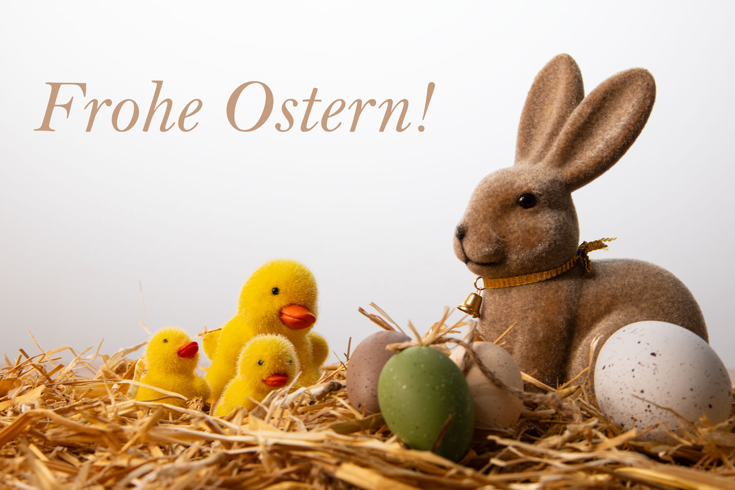 Brauner Deko-Osterhase, drei gelbe Küken und bunte Ostereier in einem Bett aus echtem Naturstroh vor einem hellen Hintergrund mit dem Text „Frohe Ostern“.