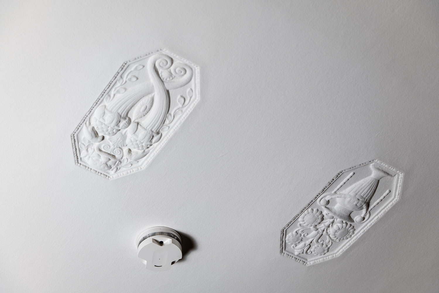 Ein moderner, weißer Rauchmelder ist an einer historischen, weißen Stuckdecke in einem Altbau montiert.